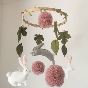 Puede incluir: Un móvil colgante con un aro de madera decorado con hojas de fieltro verde. Del aro cuelgan tres conejos de fieltro en blanco y gris, intercalados con tres pompones de lana rosa empolvado y hojas de fieltro adicionales.