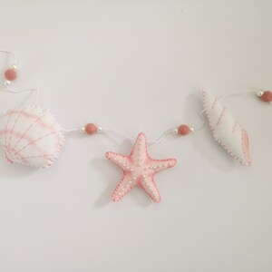 May include: A garland featuring white and pink felt sea creatures including a scallop shell, a starfish, and a spiral shell. The items are strung on a white cord interspersed with small white beads and dusty rose-colored felt balls.