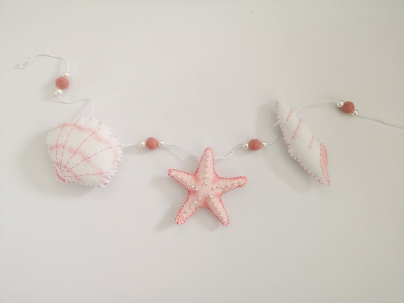May include: A garland featuring white and pink felt sea creatures including a scallop shell, a starfish, and a spiral shell. The items are strung on a white cord interspersed with small white beads and dusty rose-colored felt balls.