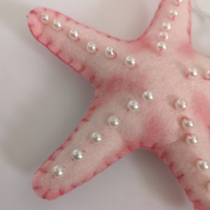 May include: A soft, light pink felt starfish ornament featuring white pearl-like beads stitched along each of its five arms. The edges are finished with visible pink thread stitching, and a small felt ball is attached to the top of the starfish.