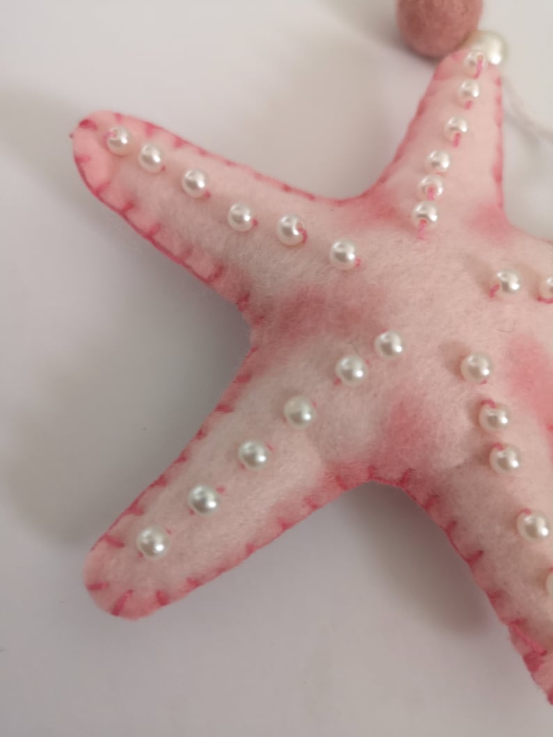 May include: A soft, light pink felt starfish ornament featuring white pearl-like beads stitched along each of its five arms. The edges are finished with visible pink thread stitching, and a small felt ball is attached to the top of the starfish.