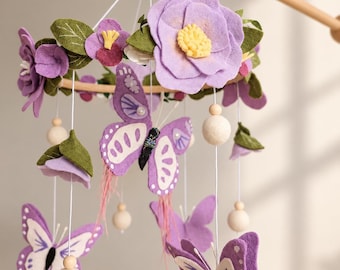 Könnte beinhalten: Ein hängendes Mobile mit Filzschmetterlingen und Blumen in Lila-, Lavendel- und Grüntönen. Der kreisförmige Holzrahmen ist mit geschichteten Filzblüten, grünen Blättern und hängenden Schmetterlingen mit weißen Akzenten verziert, die mit kleinen Holzperlen an weißen Schnüren durchsetzt sind.