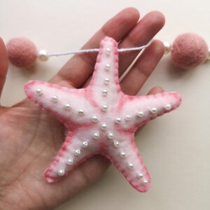 May include: A felt starfish ornament in a soft pink hue with darker pink edges, embellished with small white faux pearls. The starfish is attached to a white string featuring round, dusty pink felt balls and additional white faux pearls.