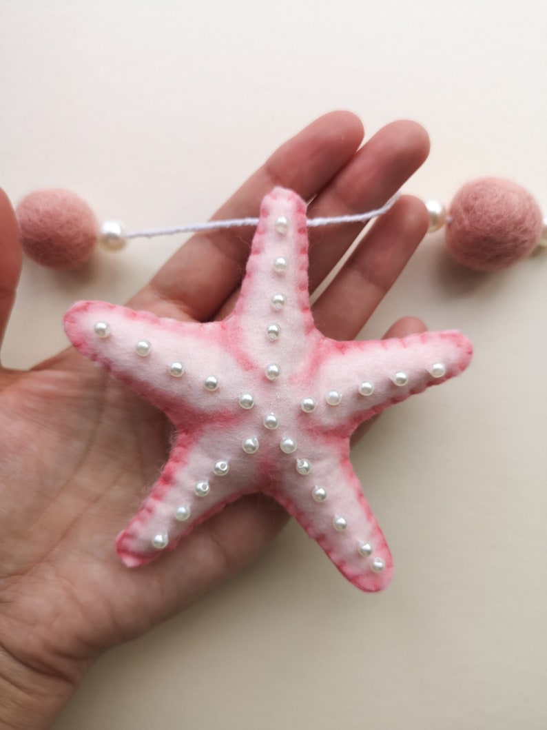 May include: A felt starfish ornament in a soft pink hue with darker pink edges, embellished with small white faux pearls. The starfish is attached to a white string featuring round, dusty pink felt balls and additional white faux pearls.