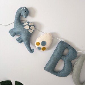 Pode incluir: Uma faixa de feltro com as letras "BOO" em azul e cinza, um dinossauro azul e um ovo branco com manchas amarelas e azuis.