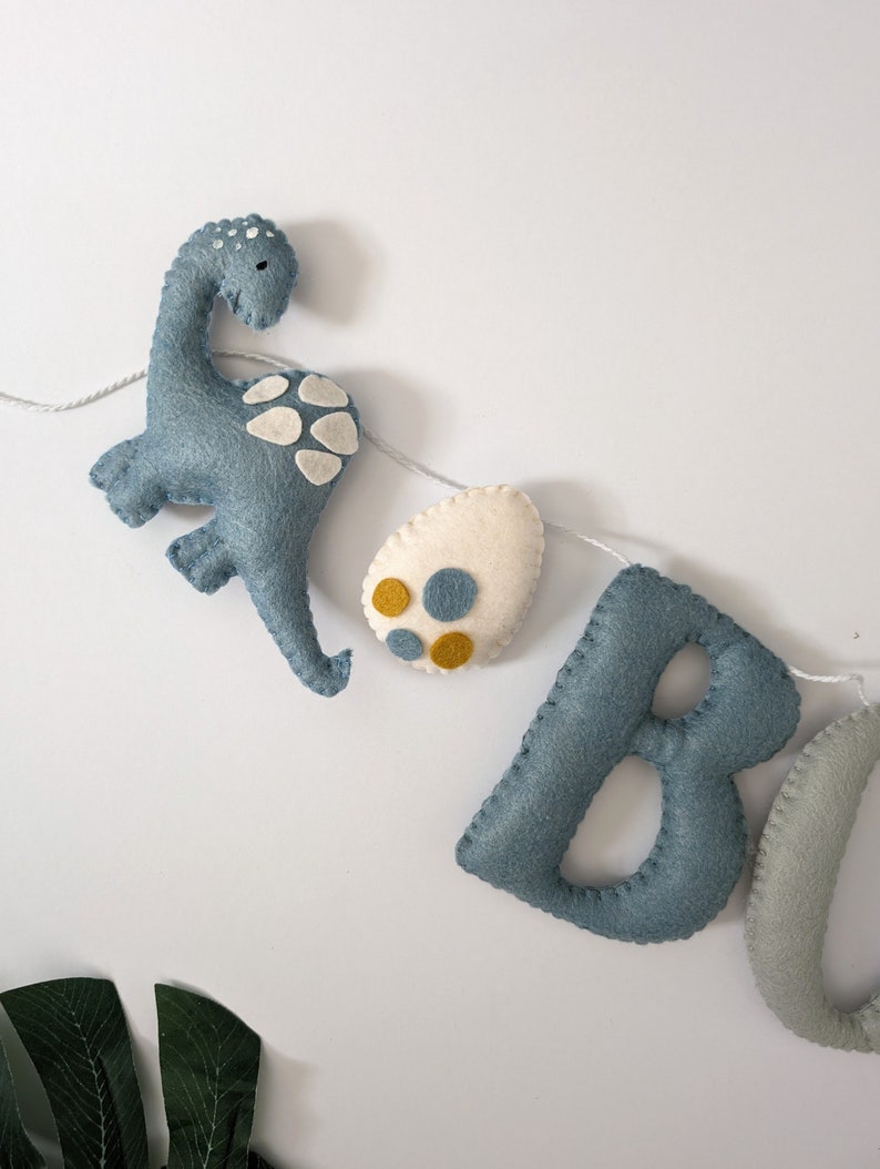 Pode incluir: Uma faixa de feltro com as letras "BOO" em azul e cinza, um dinossauro azul e um ovo branco com manchas amarelas e azuis.