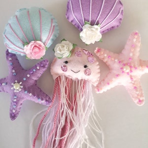 May include: A set of five felt sea-themed ornaments, including a purple starfish, a pink starfish, a light blue seashell, a purple seashell, and a pink jellyfish with a pink and white yarn tail.