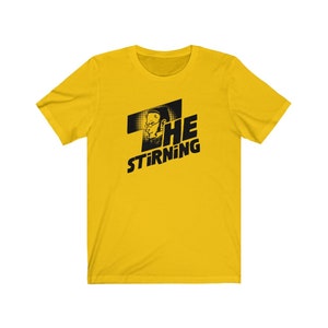 May include: Yellow short-sleeved t-shirt with a black graphic of a person's face and the text "THE STIRRING".