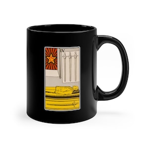 May include: Black coffee mug with a white image of a tarot card. The card is the Four of Swords, with a red star and a hammer and sickle. The card depicts a person lying down in a coffin.