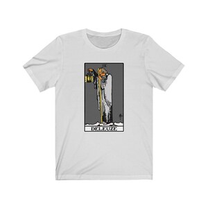 K&ouml;nnte beinhalten: Wei&szlig;es T-Shirt mit einer Schwarzwei&szlig;illustration einer Tarotkarte. Die Karte ist die 9 der St&auml;be und zeigt einen Mann, der einen Stab und eine Laterne h&auml;lt. Der Text "DELEUZE" ist unter der Karte gedruckt.
