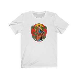 May include: White t-shirt with a colorful illustration of a woman in a robe holding a book. The illustration is surrounded by green leaves and a red circle. The text "Hildegard" is printed below the illustration.