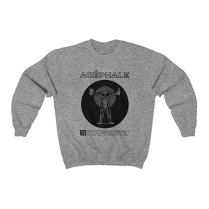 May include: A gray sweatshirt with a black circle graphic featuring a stylized human figure with the text "ACÉPHALE" above the graphic. The text below the graphic reads "TA CORRUPTION SACRÉE".