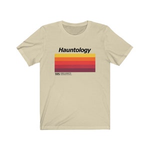 Può includere: Una t-shirt beige con un design a nastro VHS retrò. Il testo "Hauntology" è stampato in nero sul nastro. Il nastro ha uno schema di colori arcobaleno con strisce rosse, arancioni, gialle e viola.