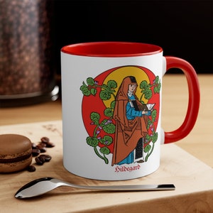 May include: White ceramic mug with a red rim and handle. The mug features a colorful illustration of a woman holding a book, surrounded by plants. The text "Hildegard" is printed at the bottom of the mug.