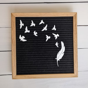 May include: A black letter board with a white feather and white paper cutouts of birds flying away from the feather. The board is framed in wood.