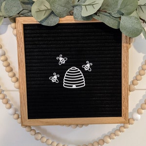 May include: A black letter board with a white honeycomb and four white bees. The letter board is framed in wood.