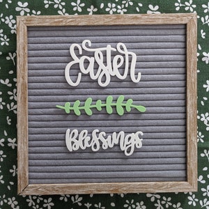 May include: A gray letter board with a wooden frame. The board has white letters spelling "Easter Blessings" with a green leaf sprig in between the words.