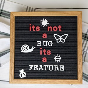 May include: A black letter board with a white wooden frame displays the message "its not a BUG its a FEATURE" in white letters with red accents.  The message is surrounded by various white insect and animal cutouts.