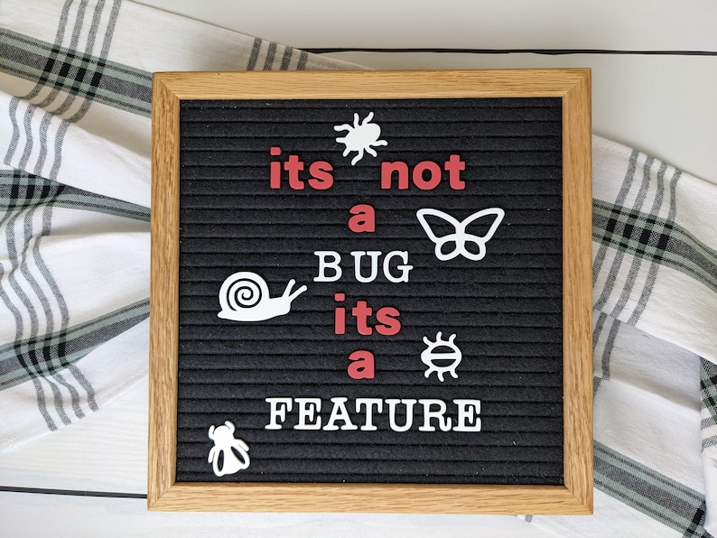 May include: A black letter board with a white wooden frame displays the message "its not a BUG its a FEATURE" in white letters with red accents.  The message is surrounded by various white insect and animal cutouts.