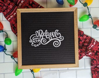 Può includere: Una lavagna nera con un ritaglio bianco di Babbo Natale e la parola "Believe" in un carattere decorativo. La lavagna è circondata da luci natalizie rosse e bianche e un tessuto a motivi rossi e bianchi.