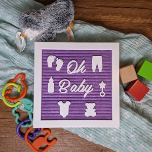 Peut inclure: Un tableau d'affichage en feutre violet avec des lettres blanches qui &eacute;pellent "Oh Baby" entour&eacute; d'ic&ocirc;nes sur le th&egrave;me du b&eacute;b&eacute;. Le tableau est sur une surface en bois avec une couverture bleue et blanche, un pingouin en peluche et des blocs en bois.
