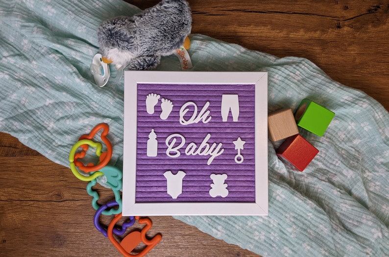 Peut inclure: Un tableau d'affichage en feutre violet avec des lettres blanches qui &eacute;pellent "Oh Baby" entour&eacute; d'ic&ocirc;nes sur le th&egrave;me du b&eacute;b&eacute;. Le tableau est sur une surface en bois avec une couverture bleue et blanche, un pingouin en peluche et des blocs en bois.