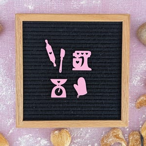 May include: A black letter board with pink felt letters spelling out "Bake with Love". The letters are arranged in a heart shape. The board is surrounded by various baked goods, including bread rolls and croissants.