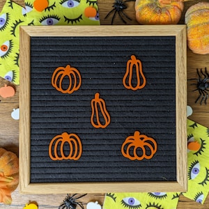 May include: A black letter board with a wooden frame, decorated with orange felt pumpkin shapes. The board is surrounded by autumn-themed decorations, including orange pumpkins, spiderwebs, and eyes.