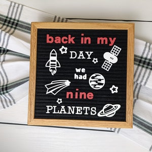 May include: A black letter board with a white wooden frame. The board has white letters spelling "back in my day we had nine planets" with white space-themed icons. The word "nine" is in red.