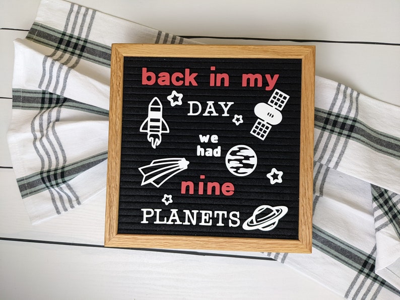 May include: A black letter board with a white wooden frame. The board has white letters spelling "back in my day we had nine planets" with white space-themed icons. The word "nine" is in red.