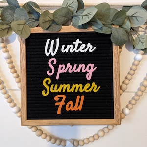 May include: A black felt letter board with a wooden frame displays the words "Winter, Spring, Summer, Fall" in colorful letters.