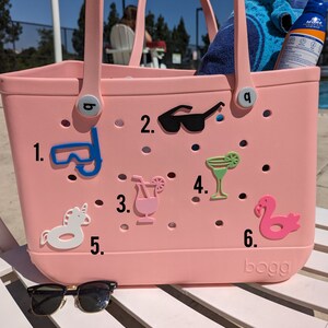 May include: A pink Bogg bag with various colorful accessories attached to it. The accessories include a pair of sunglasses, a snorkel, a cocktail, a margarita, a unicorn float, and a flamingo float. The bag has a white handle and the word "Bogg" is printed on the bottom.