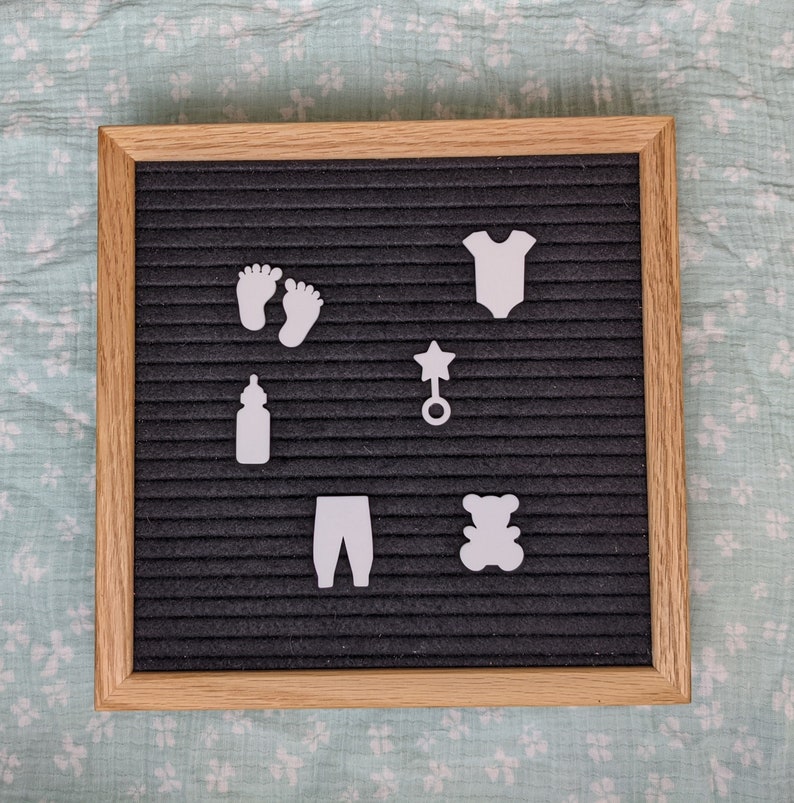 Peut inclure: Un tableau d'affichage en feutre noir avec des d&eacute;coupes en feutre blanc d'articles pour b&eacute;b&eacute;s, notamment un biberon, une grenouill&egrave;re, un hochet, une paire de pieds, un pantalon et un ours en peluche.