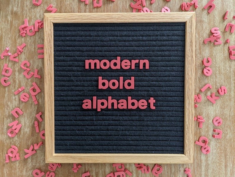 May include: A black felt letter board with a wooden frame. The board has red plastic letters spelling out "modern bold alphabet".