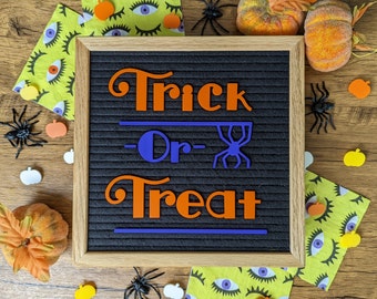 Może przedstawiać: Czarna tablica z filcu z drewnianą ramą. Tablica ma białe litery, które tworzą napis "Trick or Treat" w kolorze pomarańczowym i niebieskim. Tablica jest otoczona dekoracjami halloweenowymi, w tym pająkami, dyniami i oczami.