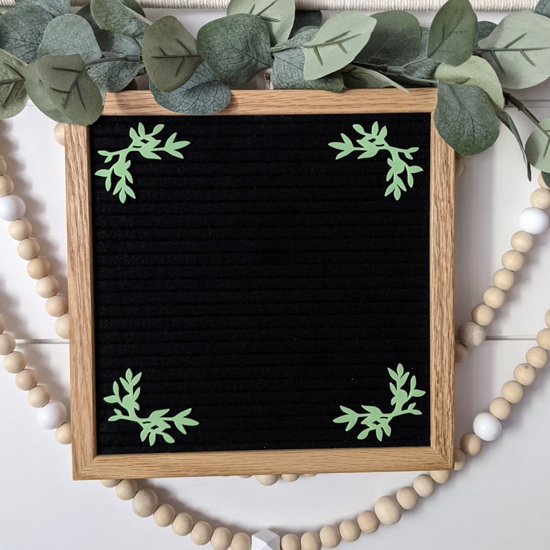 May include: A black letter board with a wooden frame. The board has green leaf-shaped decorations in each corner.