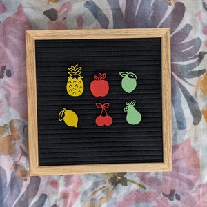 May include: A black felt letter board with yellow, green, and red felt fruit shapes. The fruit shapes include a pineapple, a lemon, a pear, an apple, and a cherry.