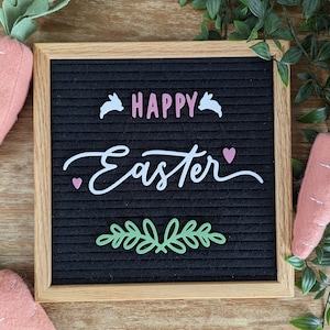 May include: A black letter board with a white wooden frame. The board has white letters spelling "Happy Easter" with pink hearts and bunny silhouettes. Green foliage and pink felt carrots are on either side of the board.
