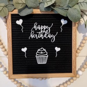 May include: A black letter board with a white wooden frame. The board has white letters spelling "happy birthday" in cursive. There are also four white heart-shaped balloons and a white cupcake illustration.
