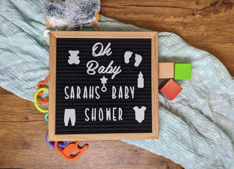 Peut inclure: Un tableau d'affichage noir avec des lettres blanches qui &eacute;pellent "Oh Baby Sarah's Baby Shower". Le tableau est d&eacute;cor&eacute; de d&eacute;coupes en feutre blanc d'un ours en peluche, d'un biberon, d'une &eacute;toile, d'une sucette et d'une paire de pieds de b&eacute;b&eacute;. Le tableau est pos&eacute; sur une surface en bois avec une couverture bleue et blanche et quelques blocs de bois color&eacute;s.