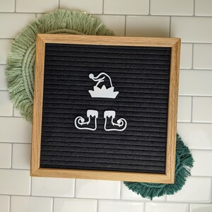 May include: A black felt letter board with a wooden frame. The board has white felt letters spelling out "Elf" with a hat and shoes.