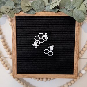 May include: Two white honeycomb shapes with bees on a black letter board. The letter board has a wooden frame.