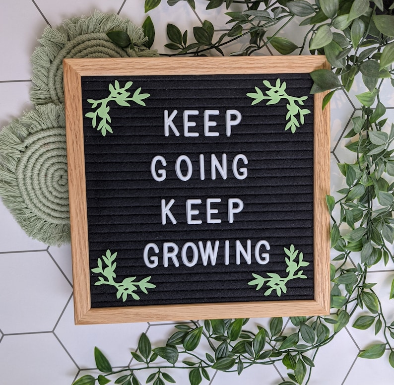 May include: A black letter board with a white wooden frame and green leaf accents. The board displays the message "KEEP GOING KEEP GROWING".