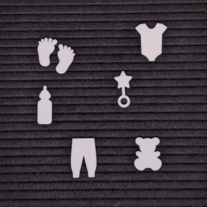 Peut inclure: D&eacute;coupes en feutre blanc d'articles pour b&eacute;b&eacute;s sur un fond noir. Les articles comprennent un biberon, une paire de chaussons, un hochet, une grenouill&egrave;re, un pantalon et un ours en peluche.