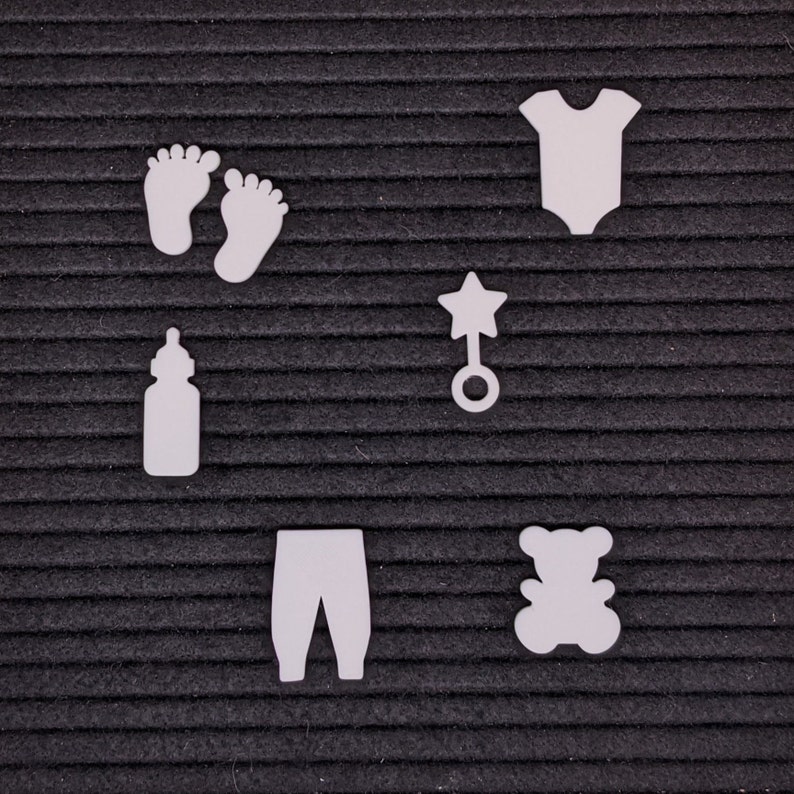 Peut inclure: D&eacute;coupes en feutre blanc d'articles pour b&eacute;b&eacute;s sur un fond noir. Les articles comprennent un biberon, une paire de chaussons, un hochet, une grenouill&egrave;re, un pantalon et un ours en peluche.