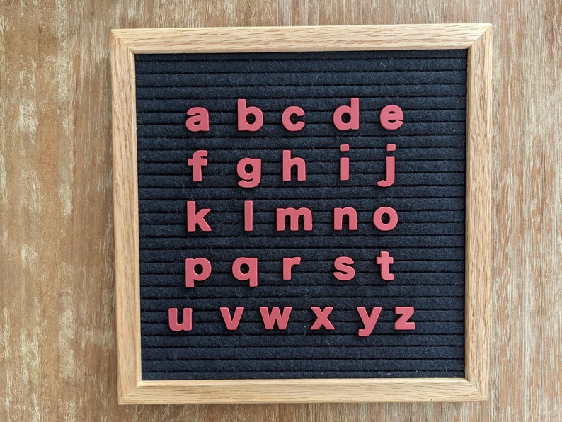May include: A black felt letter board with a wooden frame. The letters of the alphabet are displayed in red on the board.