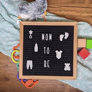 Peut inclure: Un tableau d'affichage noir avec des lettres blanches qui &eacute;pellent "MOM TO BE" et des ic&ocirc;nes blanches, notamment une &eacute;toile, un biberon, un body de b&eacute;b&eacute;, des empreintes de pieds et un ours en peluche. Le tableau d'affichage est sur une surface en bois avec une couverture bleue et blanche et des jouets color&eacute;s.
