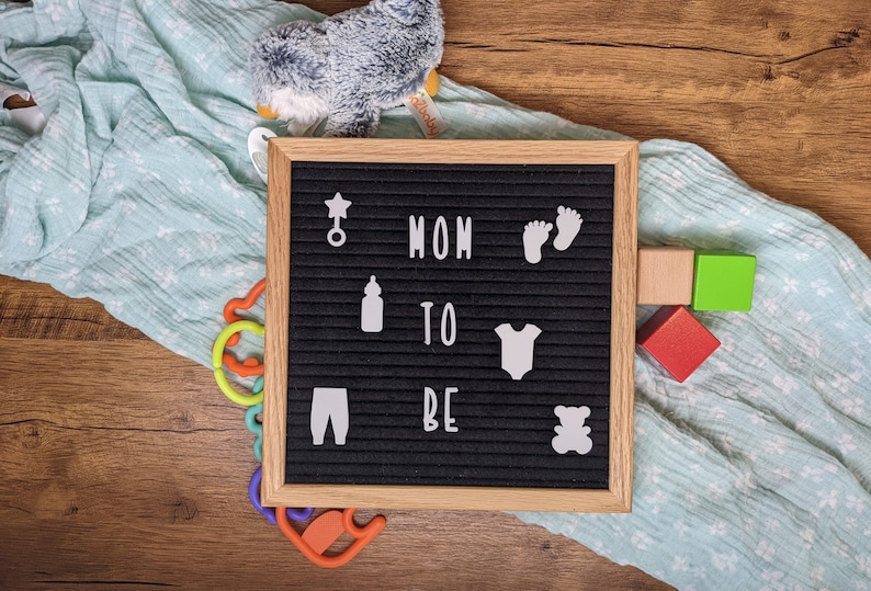 Peut inclure: Un tableau d'affichage noir avec des lettres blanches qui &eacute;pellent "MOM TO BE" et des ic&ocirc;nes blanches, notamment une &eacute;toile, un biberon, un body de b&eacute;b&eacute;, des empreintes de pieds et un ours en peluche. Le tableau d'affichage est sur une surface en bois avec une couverture bleue et blanche et des jouets color&eacute;s.