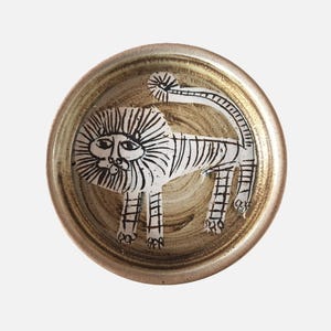 May include: A circular ceramic plate featuring a hand-drawn lion design. The lion is white with black detailing, set against a beige background with a golden rim. The lion has a detailed mane and tail.