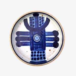 以下が含まれることがあります： 青と白の模様が施された、小さな丸い陶器の皿。デザインは、中央に円があり、そこから放射状に線が伸びた、様式化された人物を表しています。